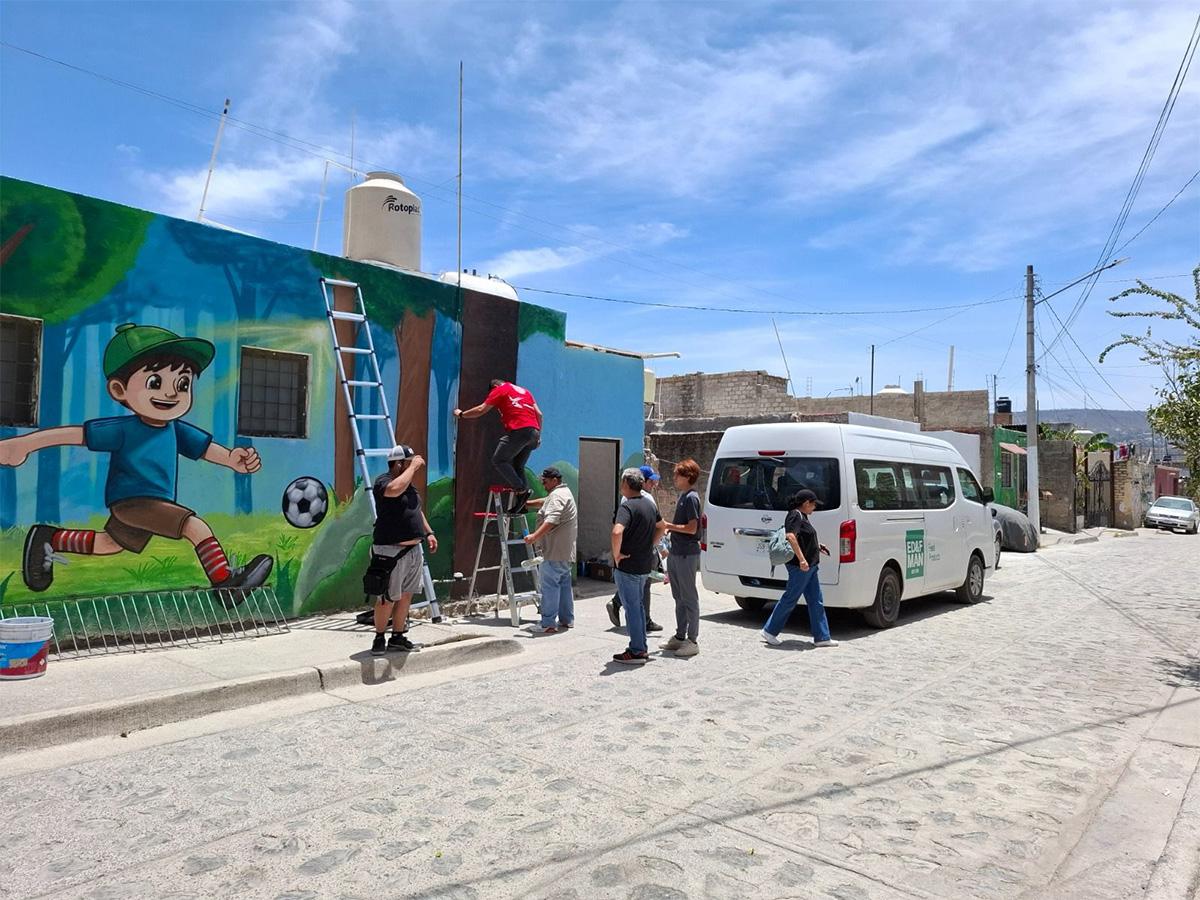 group of people painting a colorful mural of a boy kicking a soccer ball on a sunny street, with a ladder and a white van nearby.